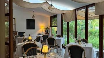 Un comedor con mesas dispuestas para dos, con manteles blancos, sillas de mimbre y una mesa central con una lámpara.