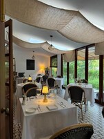 Un comedor con mesas dispuestas para dos, con manteles blancos, sillas de mimbre y una mesa central con una lámpara.