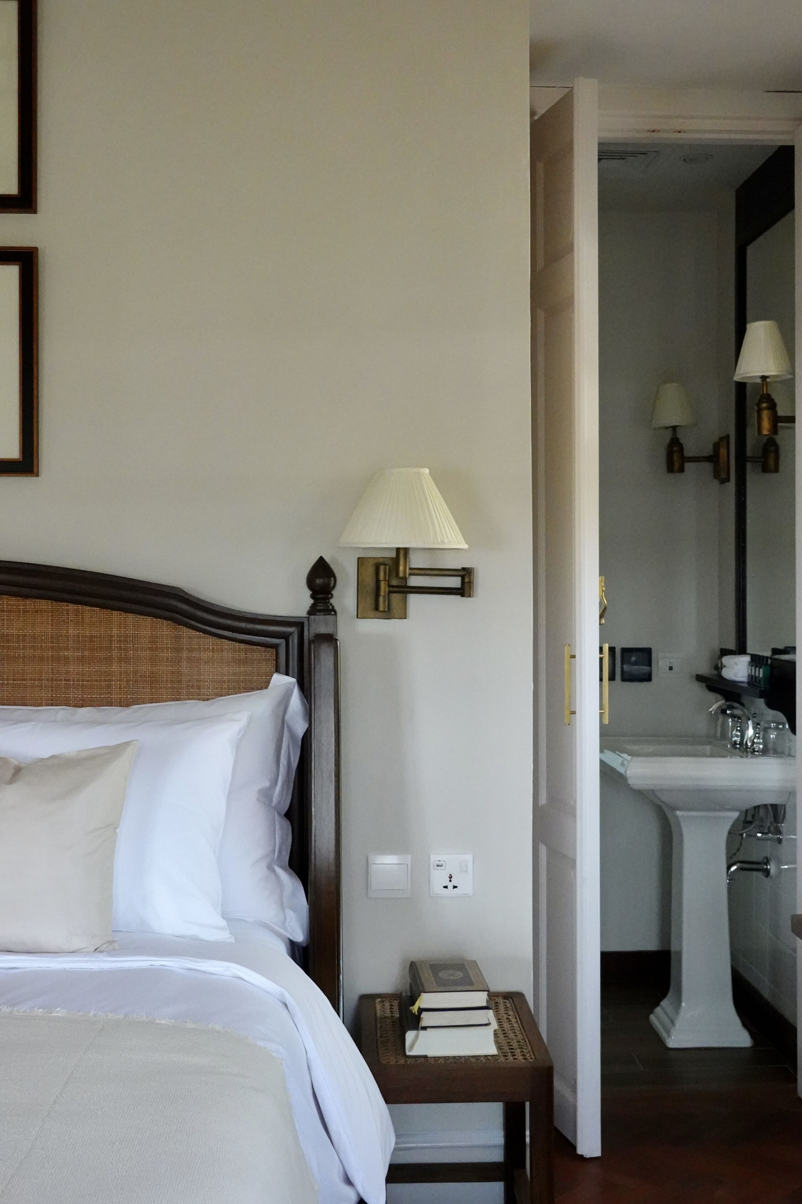 Habitación de hotel con cama, lámpara de noche, mesita de noche con libros y baño visible a través de una puerta abierta.