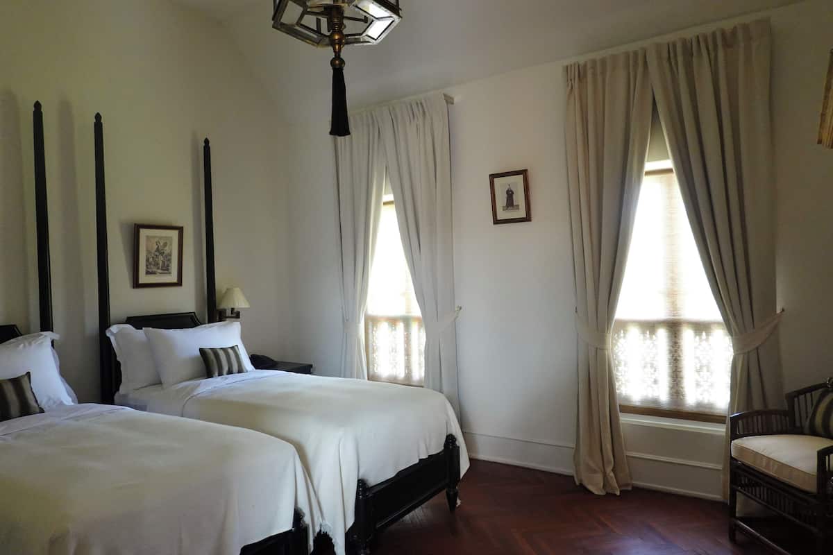 Habitación de hotel con dos camas, piso de madera, candelabro y ventanas amplias con cortinas.