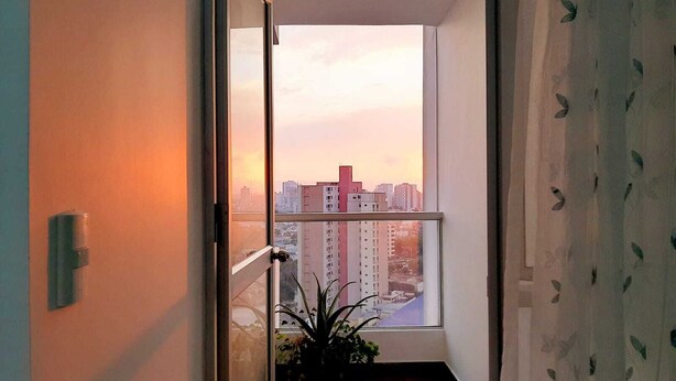 Ein schmaler Korridor mit einem großen Fenster, einem Topfpflanzen und Blick auf eine Stadtlandschaft bei Sonnenuntergang.