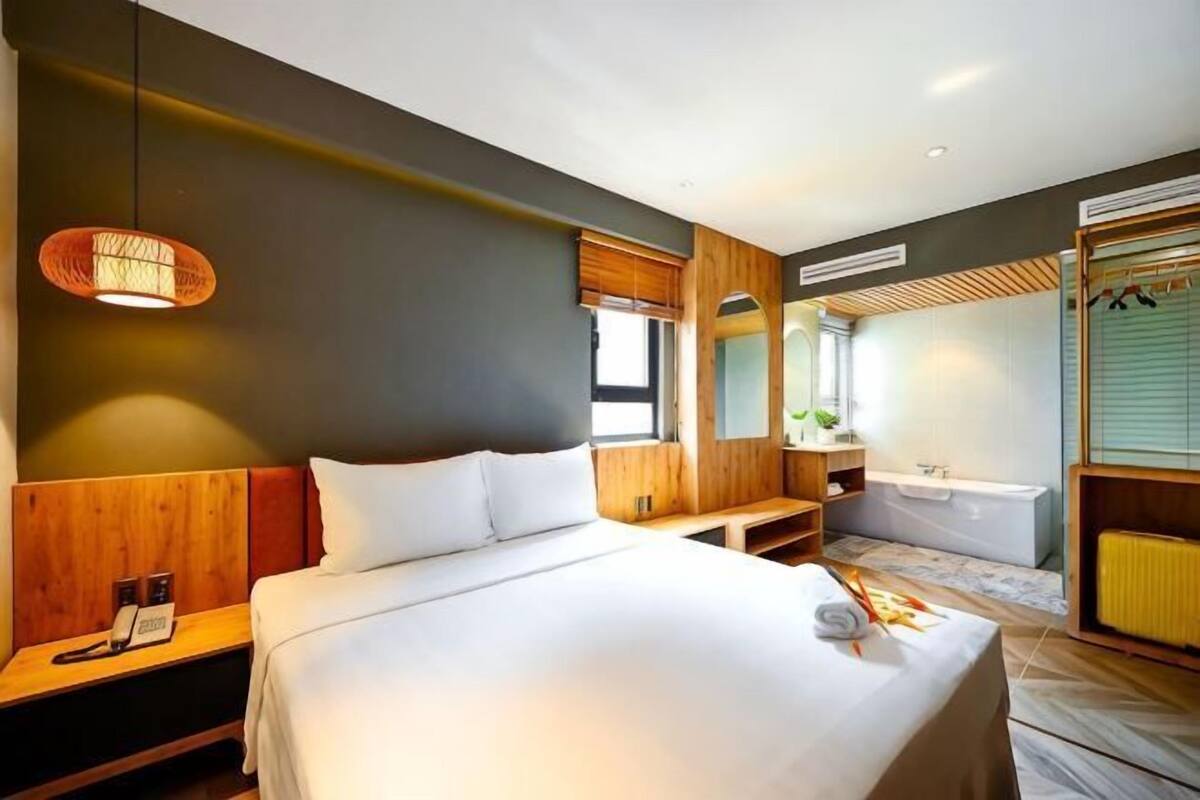 Une chambre d’hôtel moderne dotée d’un grand lit, d’une table de chevet, d’une commode, d’une salle de bain avec baignoire et d’une fenêtre avec stores.