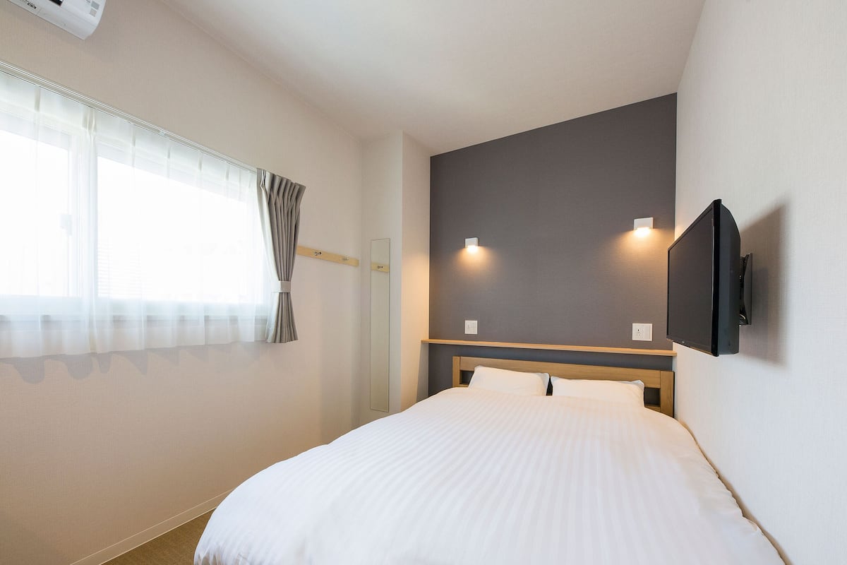 Una habitación de hotel con cama, ventana con cortinas, televisor de pared y un interruptor de luz.