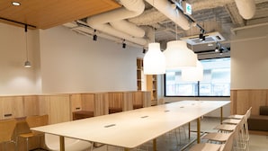 Uma sala de conferências moderna com uma mesa comprida, cadeiras brancas, luminárias pendentes e vista para a cidade através de grandes janelas.