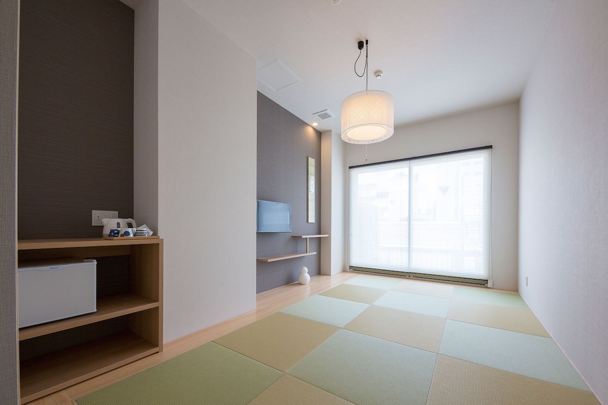 Habitación moderna y minimalista con esteras de tatami, un estante de madera bajo, un armario blanco pequeño y un ventanal con persianas.