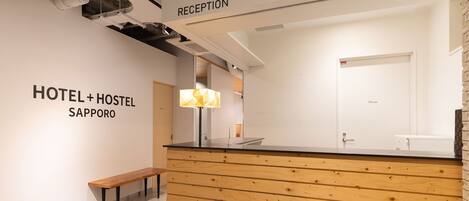 Un mostrador de recepción con diseño de panel de madera, un banco y un cartel en la pared que indica “RECEPTION” y “HOTEL + HOSTEL SAPPORO”.