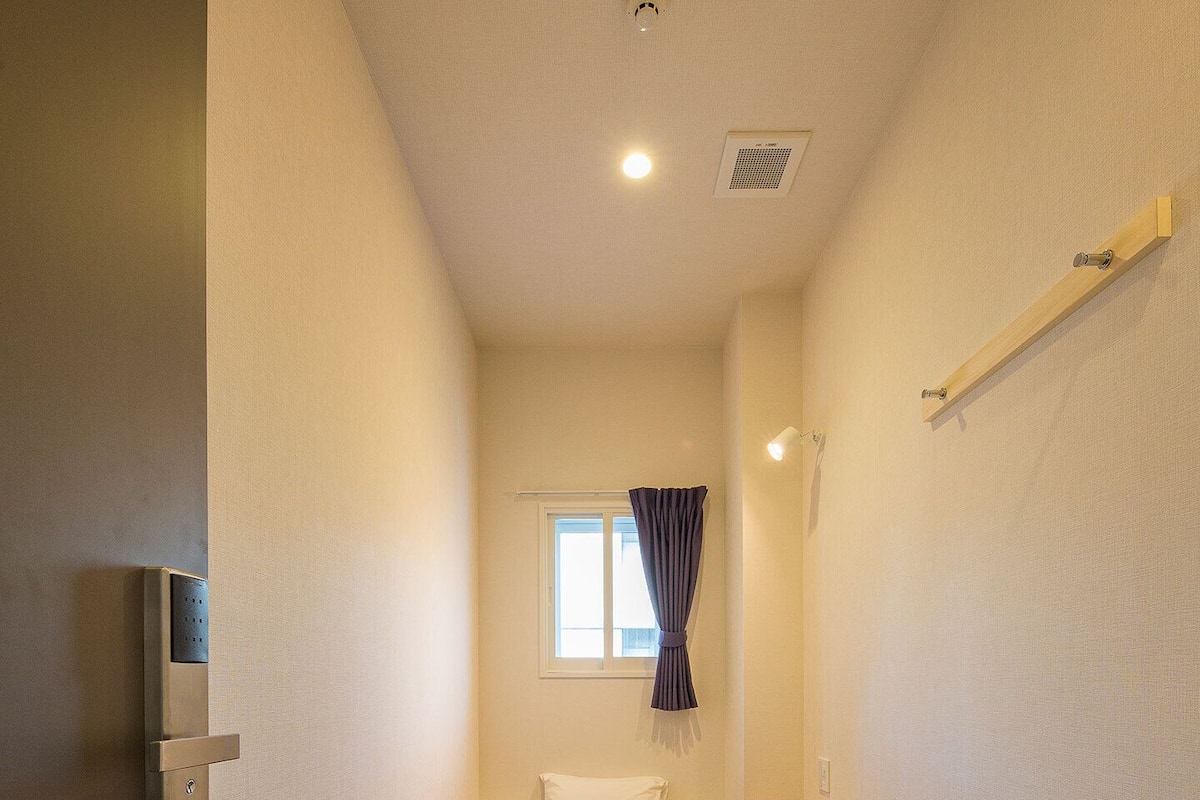 Una habitación pequeña y estrecha con una cama individual, una ventana con cortinas, una mesita de noche y un aplique de pared.
