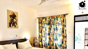A hotel room with a bed, a wall-mounted artwork, a patterned curtain, and a wicker chair. - Shanthi Boutique Villa (Hikkaduwa)