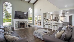 Una sala de estar amplia con chimenea, televisor grande y comedor, con una cocina al fondo.