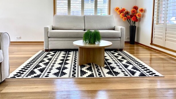 Un salon comprenant un canapé, une table basse ronde en bois, un tapis à motifs et une fenêtre avec des stores.