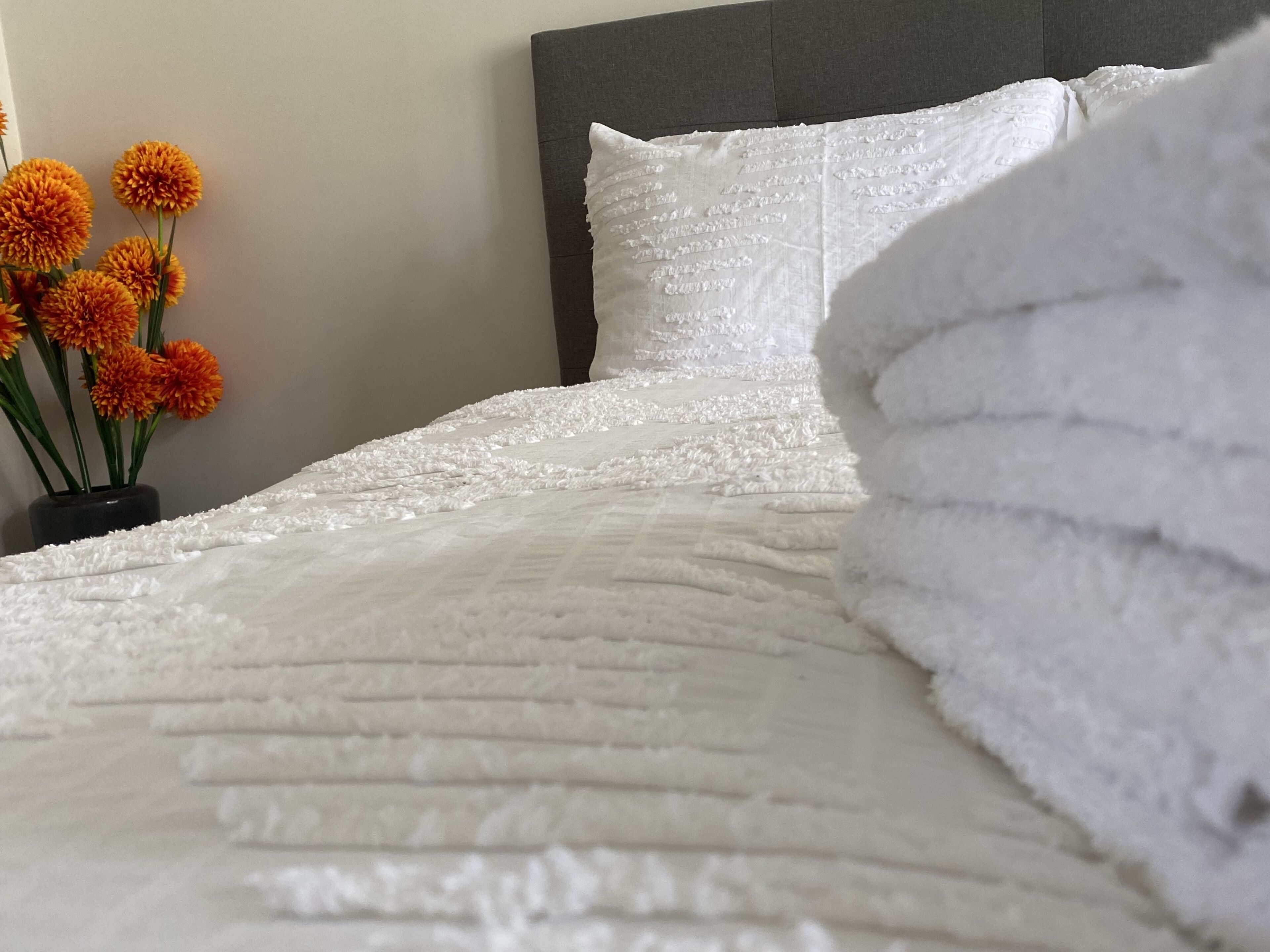 Een net opgemaakt bed met een witte quilt en kussens, een vaas met oranje bloemen, en een grijs hoofdeinde.