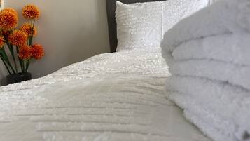 Een net opgemaakt bed met een witte quilt en kussens, een vaas met oranje bloemen, en een grijs hoofdeinde.