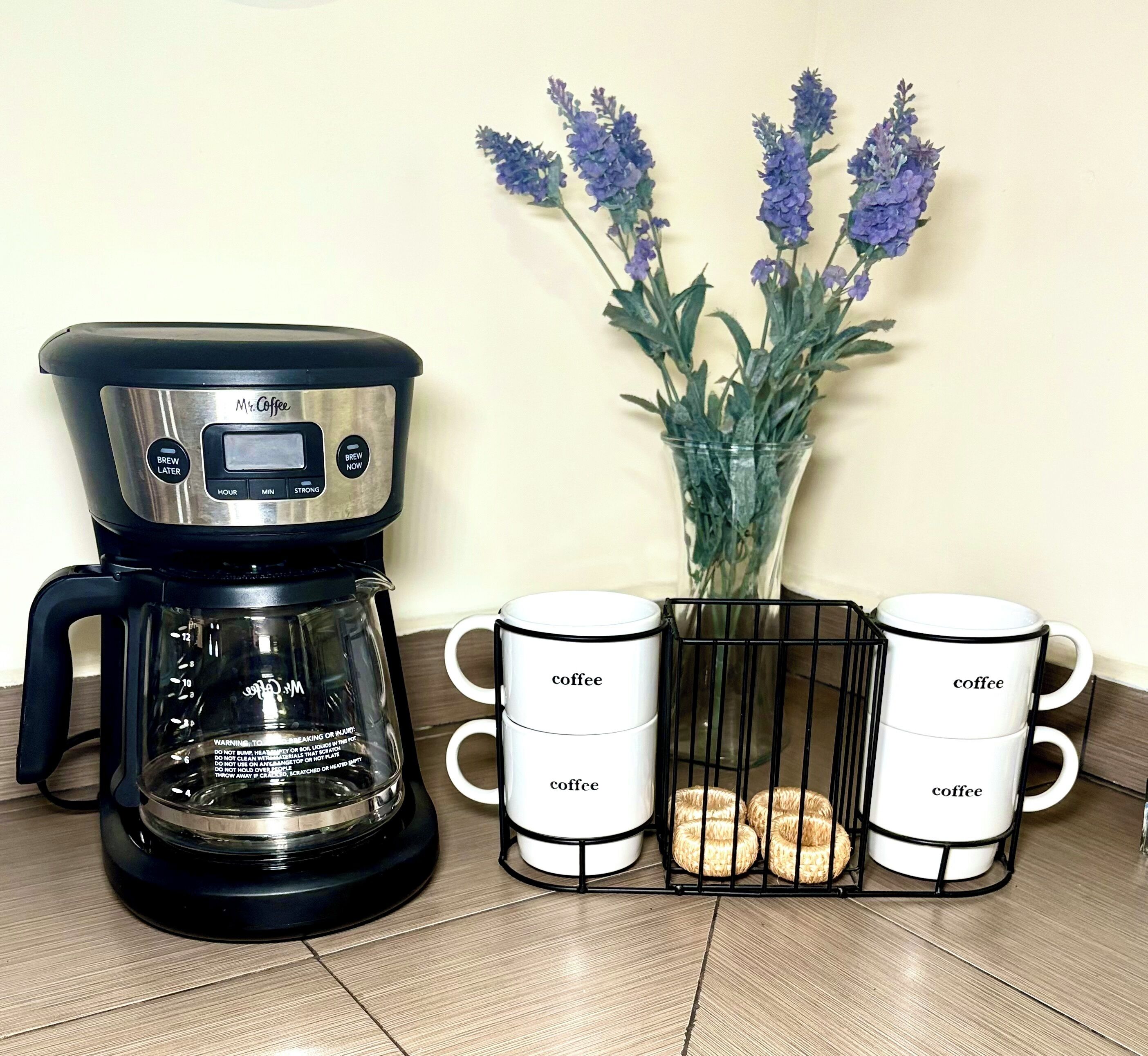 A coffee maker, two coffee mugs, and a vase with lavender flowers.