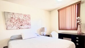 Una cama bien hecha con edredón blanco, una mesita de noche con lámpara, un cuadro colgado en la pared y una ventana con persianas.