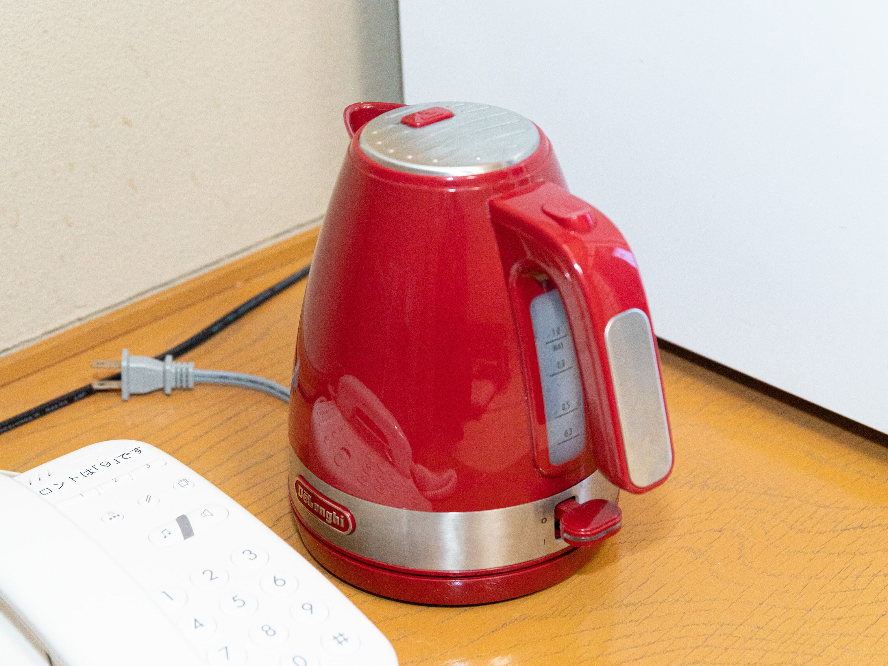 A red electric kettle on a wooden surface next to a white telephone.