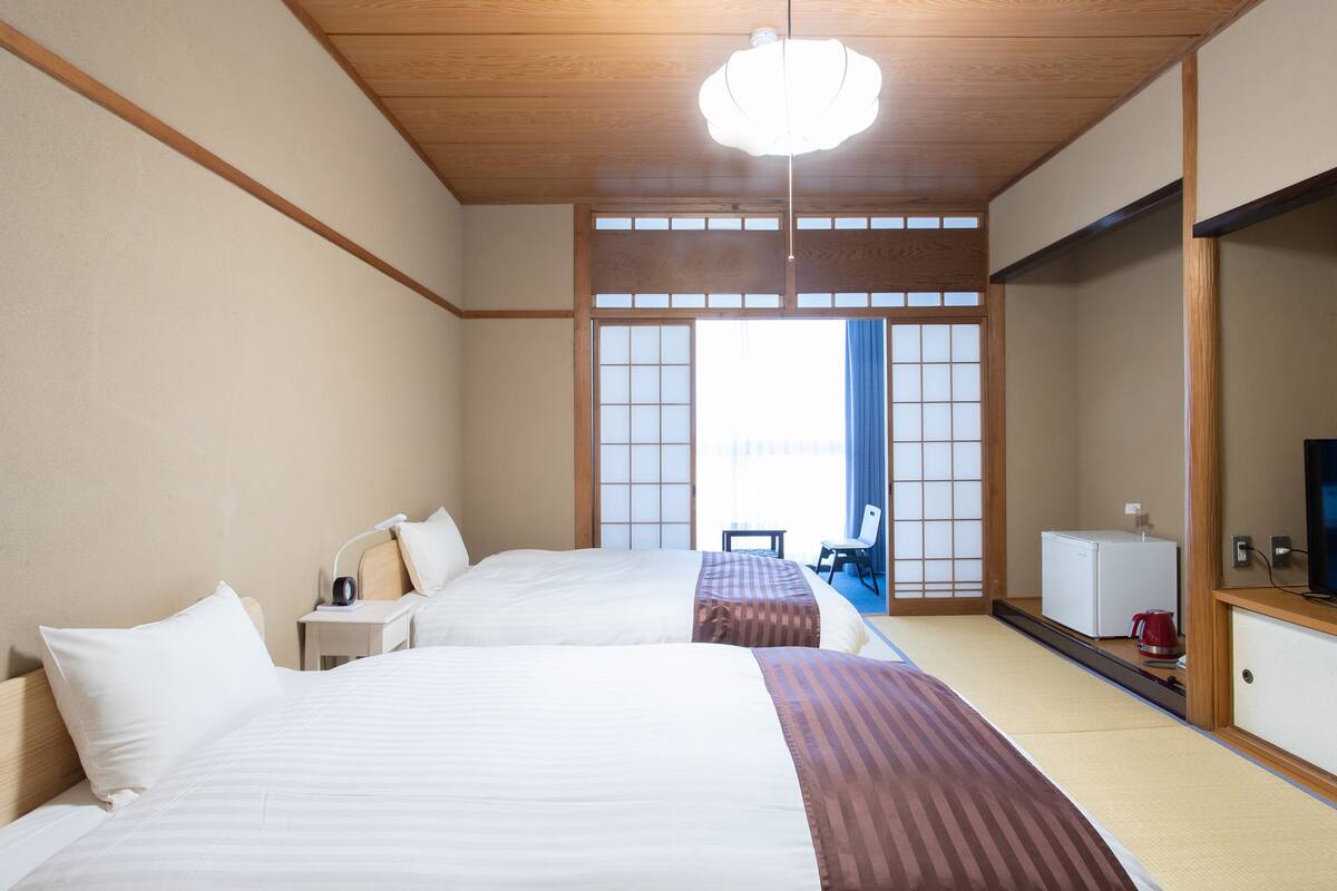 Een traditionele Japanse hotelkamer met twee bedden, een kleine tafel, een stoel, een televisie en een koelkast.