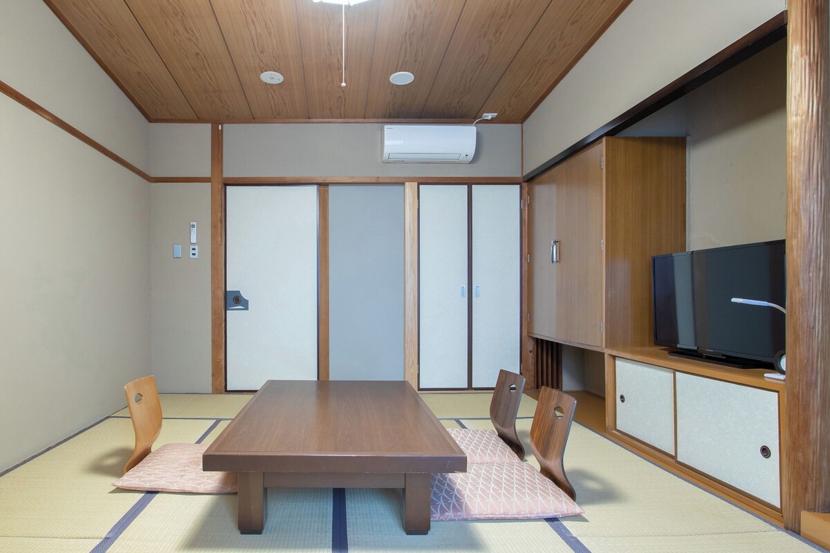 Una habitación japonesa tradicional con suelo de tatami, una mesa de madera, sillas, un televisor y puertas correderas.