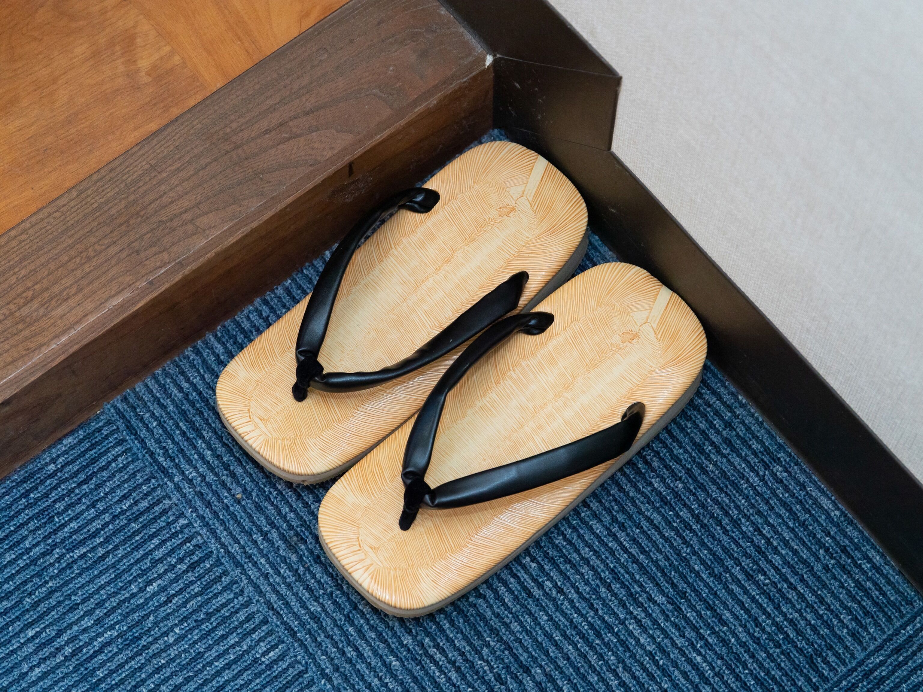 Un par de chanclas sobre un tapete azul con textura, cerca de un escalón de madera.