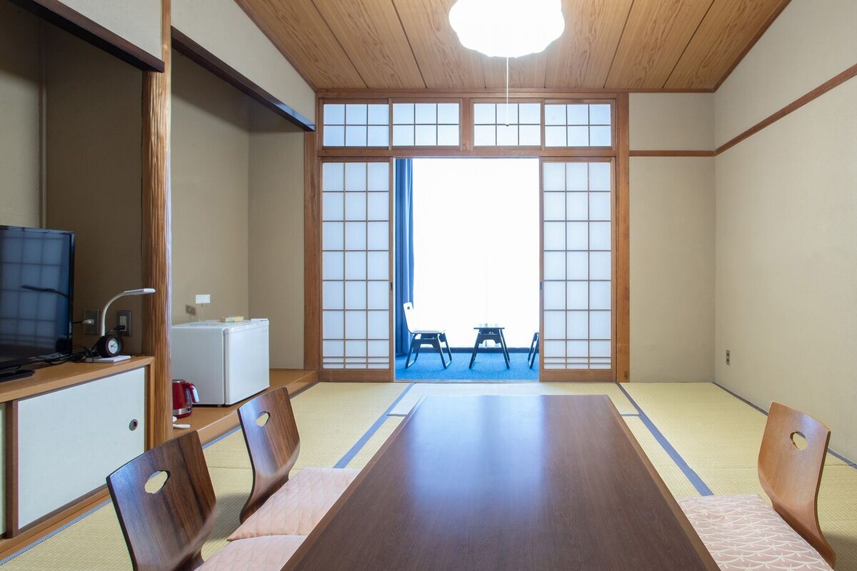 Una habitación tradicional japonesa con suelo de tatami, una mesa de madera baja y un televisor sobre un mueble.