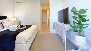 Habitación de hotel con cama, televisor, planta en maceta y un pasillo que conduce a otra habitación.