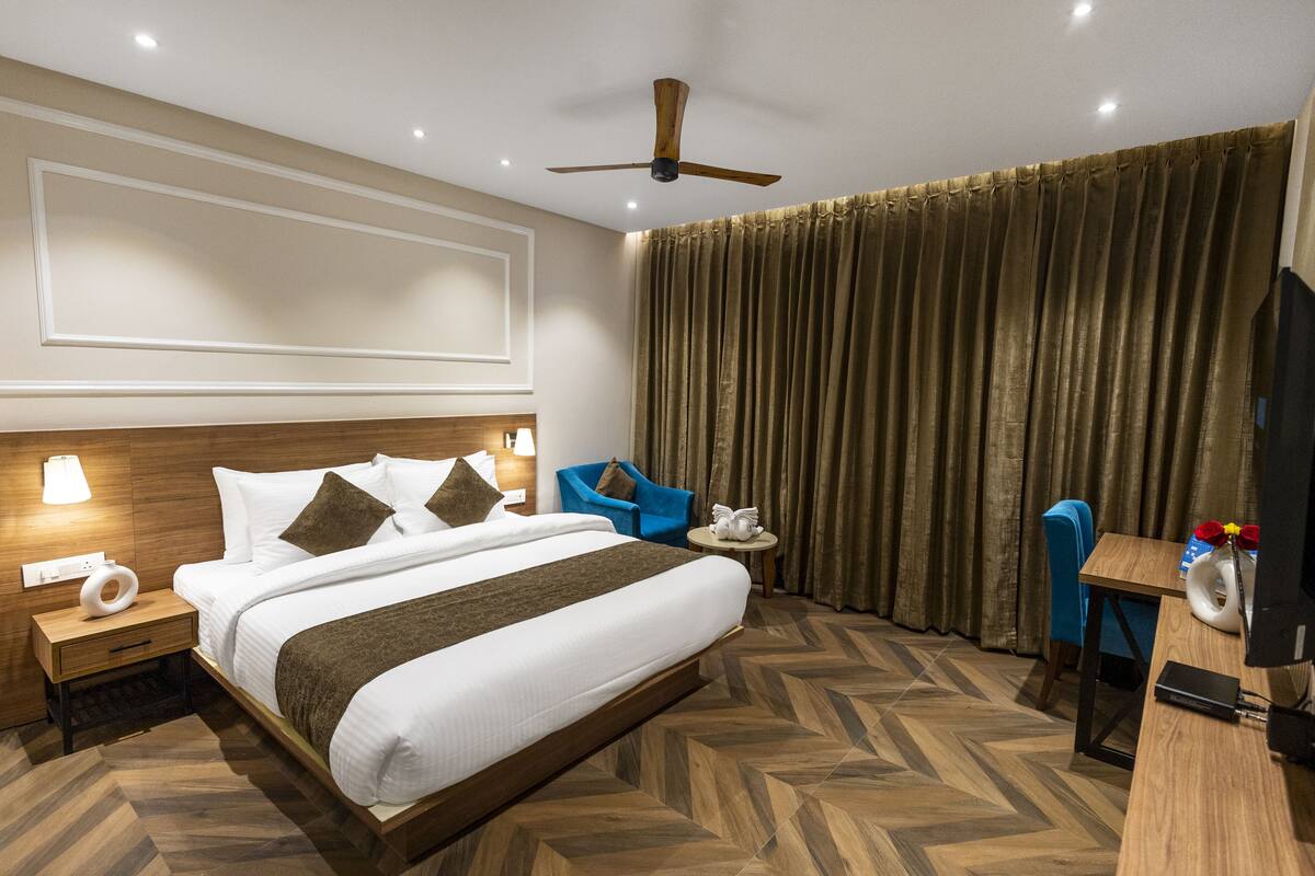 Habitación de hotel con una cama grande, cabecera de madera, ventilador de techo, escritorio con silla, televisor y ventana con cortinas.