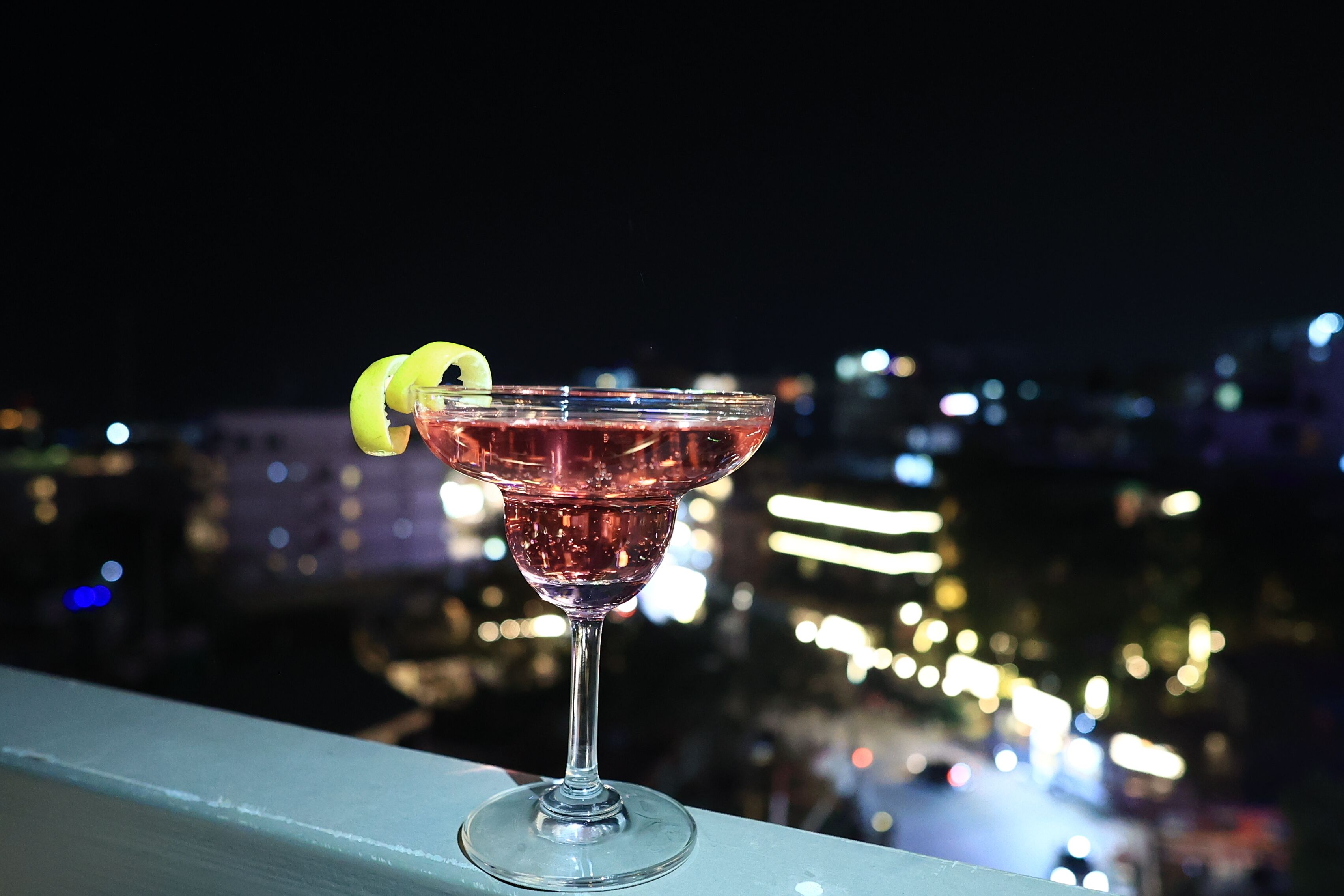 Ein Cocktailglas mit Zitronenzeste auf einem Balkon mit Blick auf eine nächtliche Skyline.