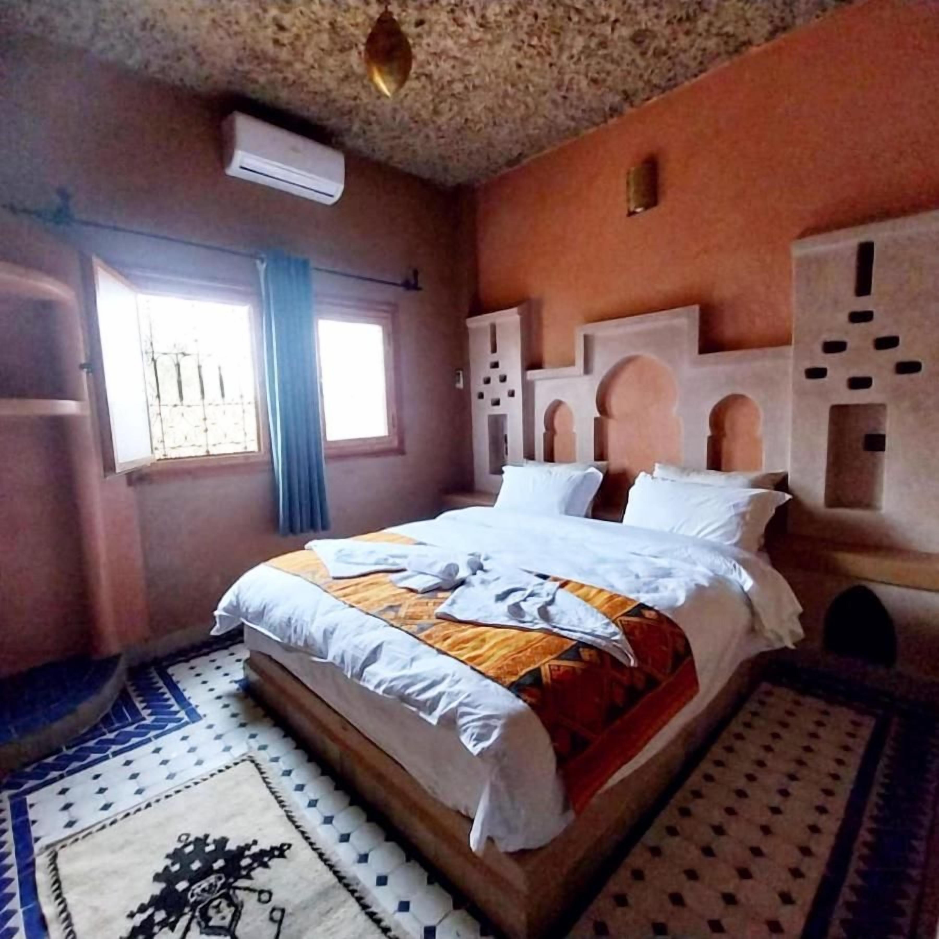 Una habitación con cama, ventana con cortinas, equipo de aire acondicionado y alfombras estampadas.