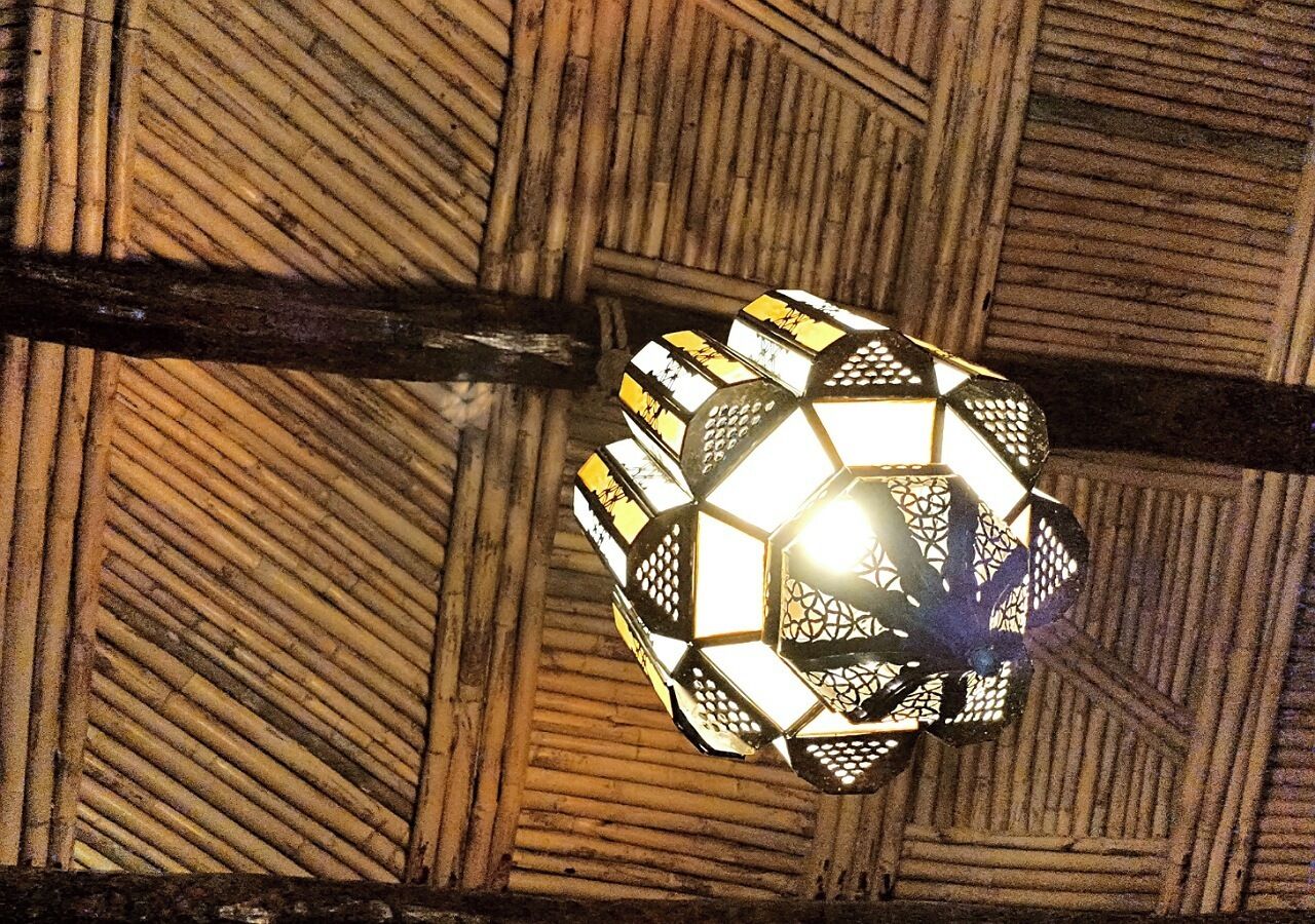 A hexagonal lantern with a geometric pattern, hanging from a wooden ceiling with a diagonal plank structure.