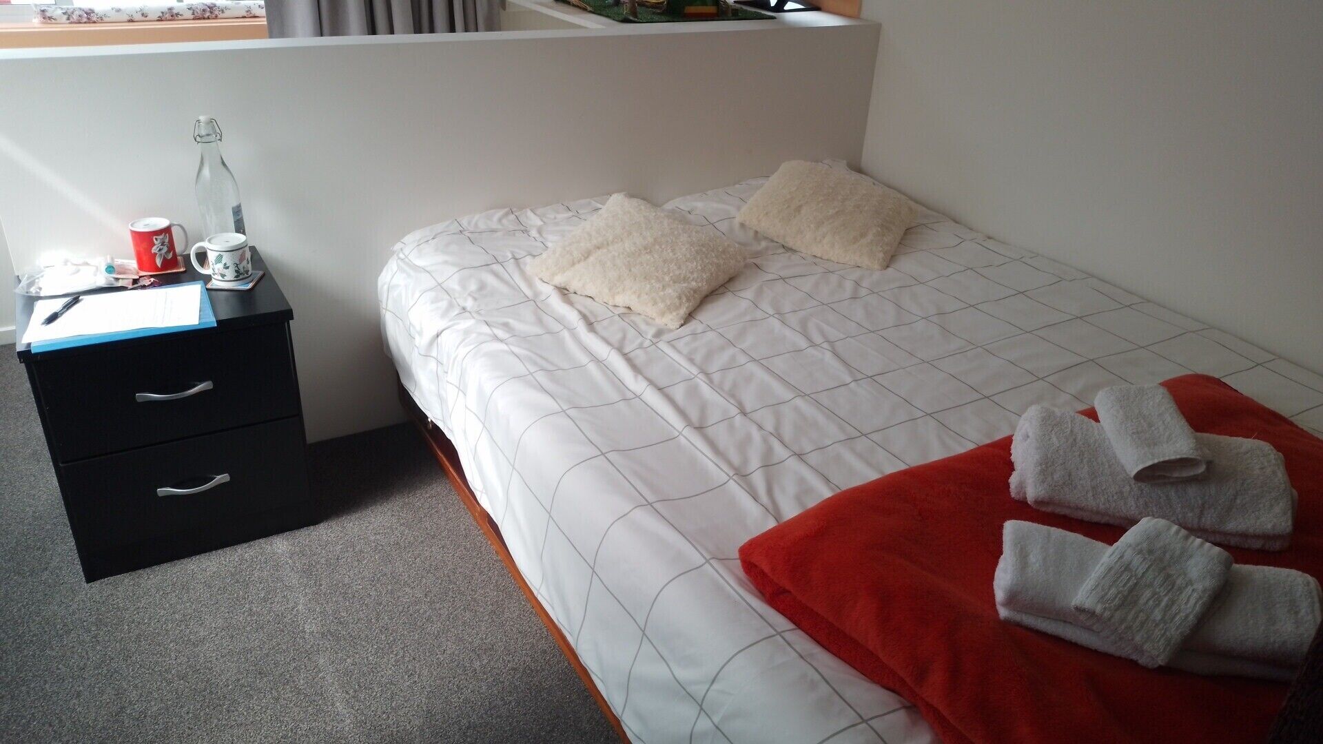 A hotel room with a bed, two pillows, a red blanket, a set of towels, a desk with a bottle, a cup, and a notebook.