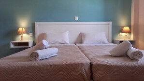 A hotel room with two beds, each with a beige quilted bedspread and white pillows. There are two bedside tables with lamps, and a light switch on the wall. - Amelia's Garden (Corfu)