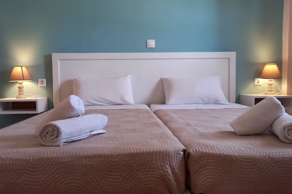 A hotel room with two beds, each with a beige quilted bedspread and white pillows. There are two bedside tables with lamps, and a light switch on the wall.