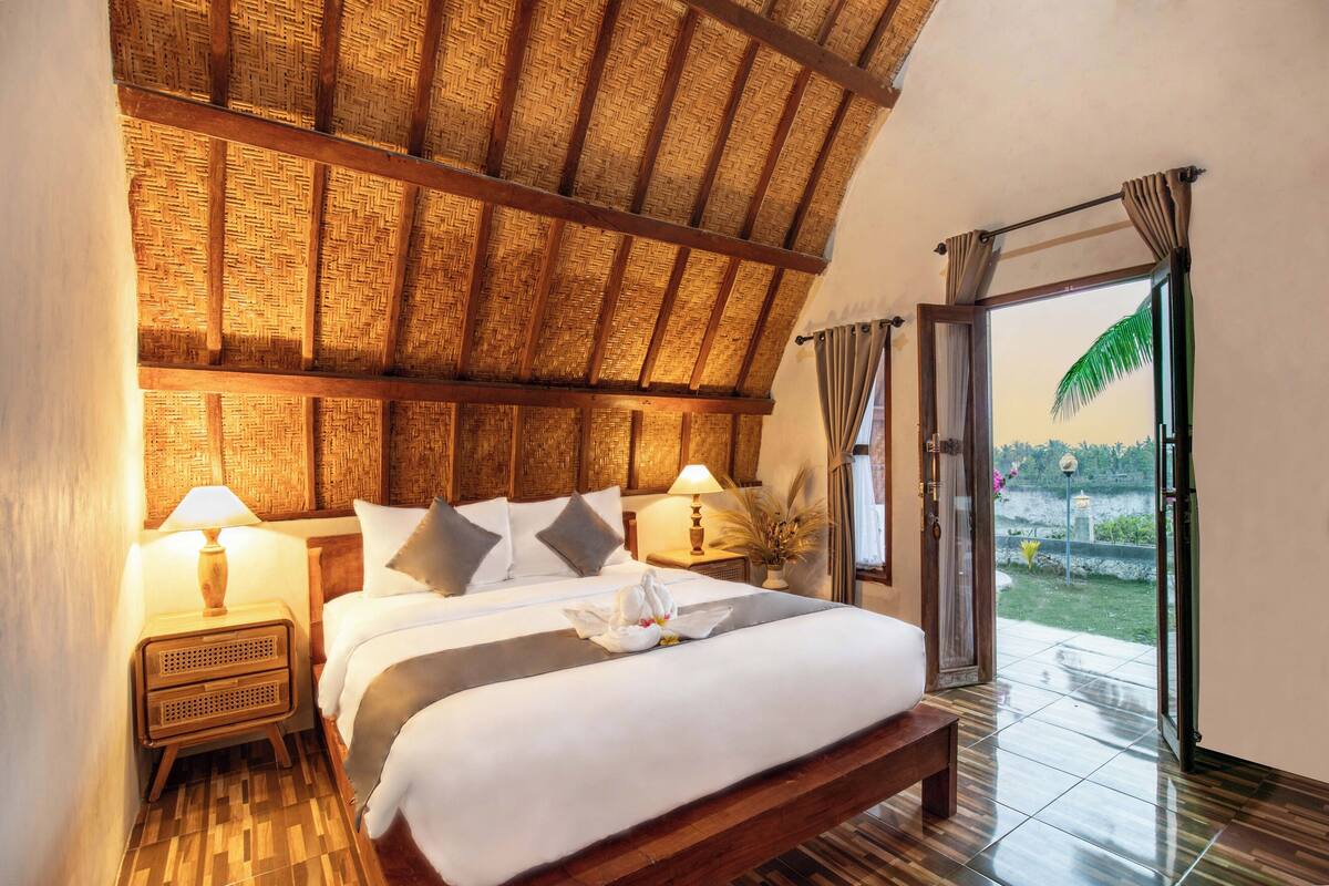 A room with a bed, nightstands, a lamp, a window with curtains, and a view of the outdoors.
