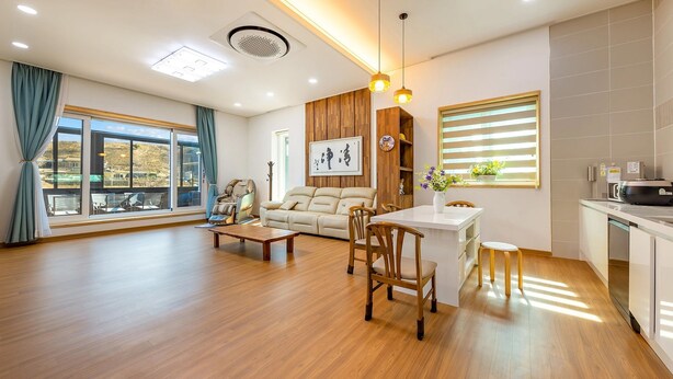 A spacious living area with a sofa, a dining table with chairs, a massage chair, and a view through large windows. - Chungnam Cheong-yang Ekohillinghauseu (Cheongyang)
