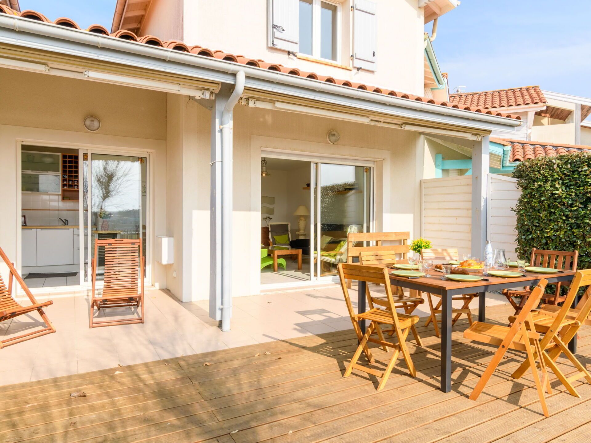 Une terrasse couverte avec une table et des chaises en bois, une porte-fenêtre coulissante donnant sur un espace de vie et un toit recouvert de tuiles.