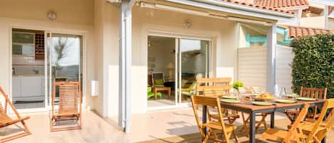 A covered patio with a wooden table and chairs, a sliding glass door leading to a living area, and a tiled roof.