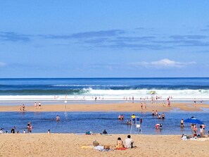 Ein Strand mit badenden und sonnenden Menschen, und ein Rettungsturm in der Ferne.