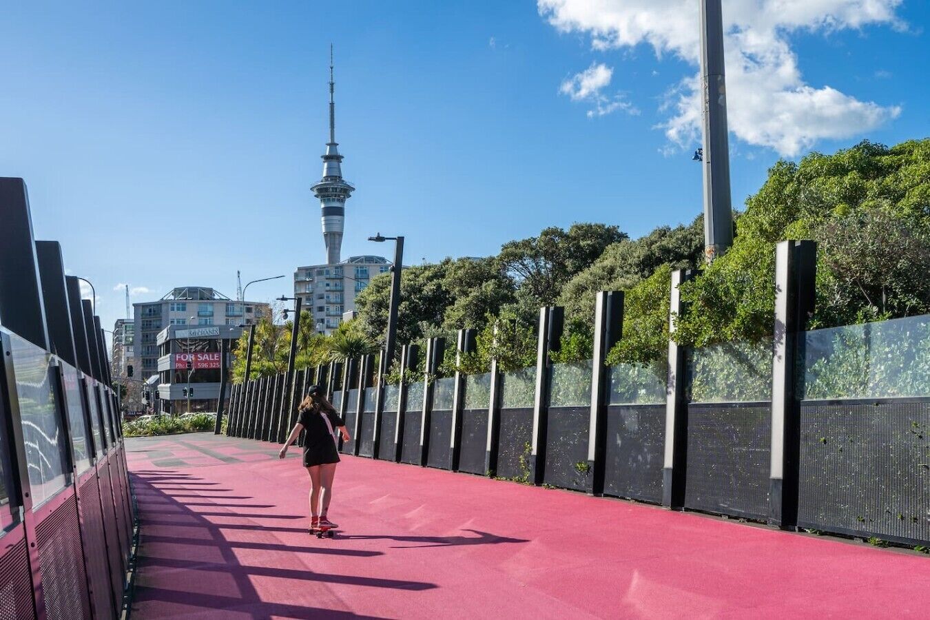 A red pedestrian pathway with a person skateboarding, a fence, and a tower in the background.