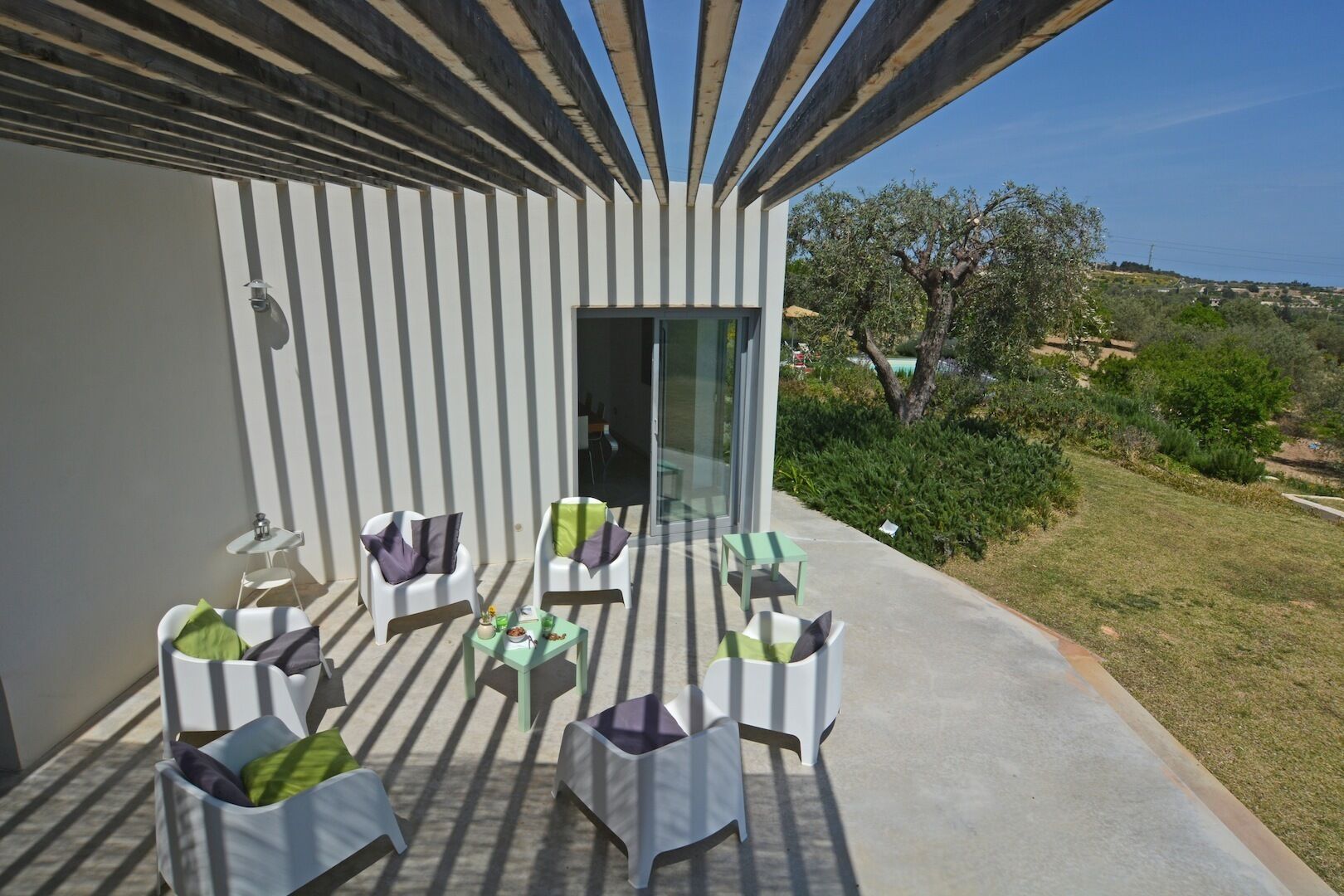 A modern outdoor patio with white chairs and tables, a glass door leading inside, and a view of the surrounding landscape.