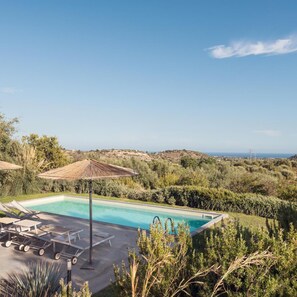 A rectangular swimming pool with lounge chairs and a table, surrounded by greenery and overlooking a hilly landscape. - Villa Cavaliere in Noto (Noto)