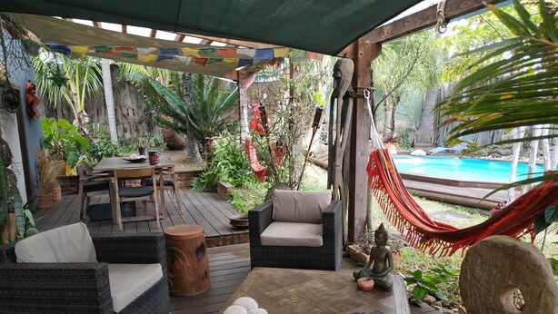 A covered outdoor area with a wooden deck, wicker furniture, a table with cushions, a small Buddha statue, and a hammock. - BAS DE VILLA cosy, wooded garden with swimming pool, very quiet, town center. (Nouméa)