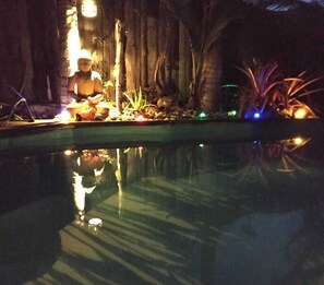 Área de piscina à noite com estátua, plantas e iluminação ambiente.