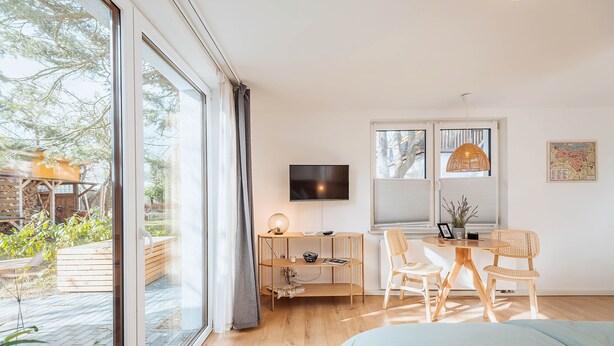 A modern room with a large sliding glass door, a flat-screen TV, a wooden desk with a lamp, two wicker chairs, and a bed with a light blue cover. - Apartments Kassebohmer Weg (Rostock)