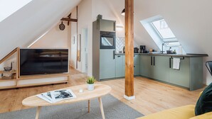 A modern loft apartment with a large TV, a dining area, a kitchen with a window, and a wooden ceiling beam. - Apartments Kassebohmer Weg (Rostock)