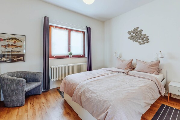 A bedroom with a bed, a chair, a window with curtains, a radiator, and wall art.