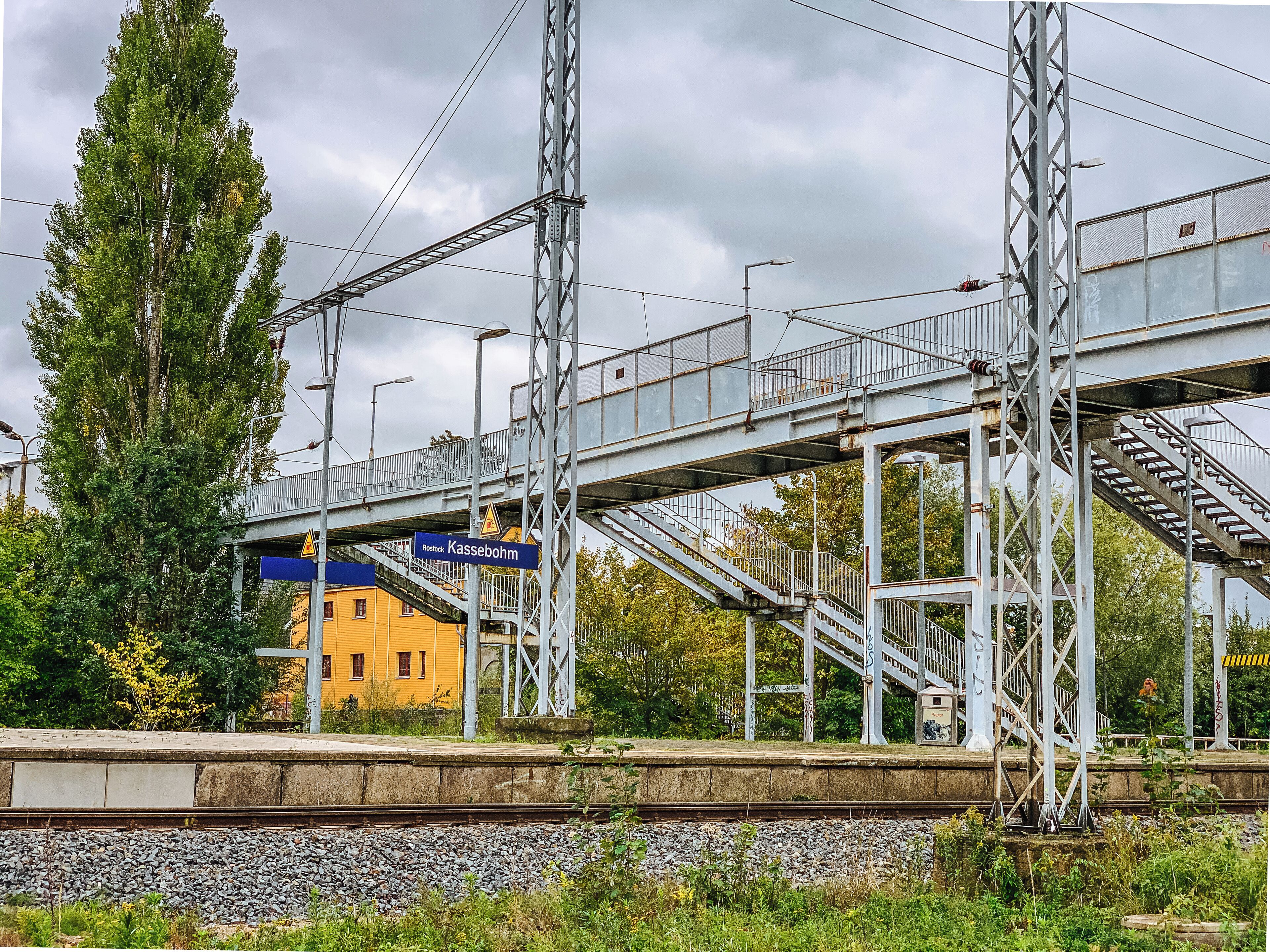 Un quai de gare avec un pont piétonnier, un bâtiment jaune et un grand arbre.