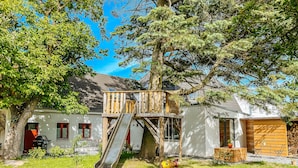 A wooden play structure with a slide, a tree, and a house with a red door and windows. - Apartments Kassebohmer Weg (Rostock)