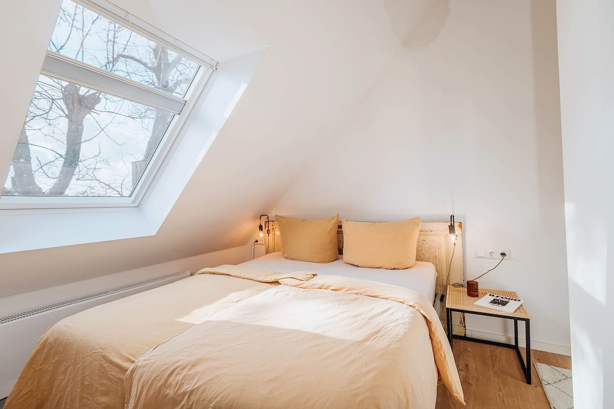 A bedroom with a skylight, a bed with beige bedding, a bedside table with a lamp, and a power outlet.