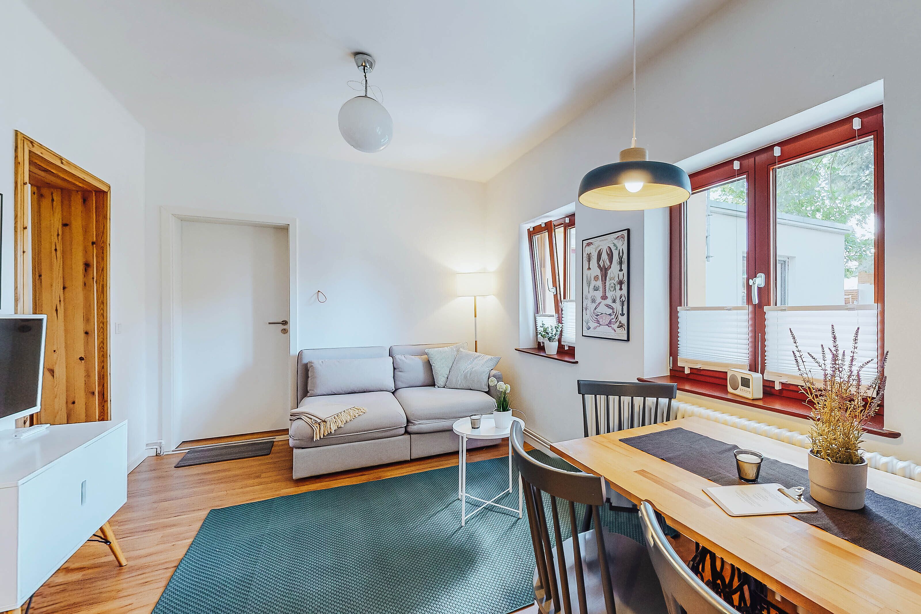 Una sala de estar moderna con un sofá, una mesa de centro y una mesa de comedor. Hay un televisor sobre un mueble blanco, una lámpara y una ventana con persianas.