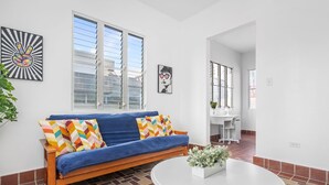 A room with a blue sofa, a white table, and a window with blinds. - Otium Rentals Plush King Bed Pacman Arcade (San Juan)