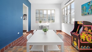 A dining area with a white table and chairs, a potted plant, and a colorful arcade machine. - Otium Rentals Plush King Bed Pacman Arcade (San Juan)